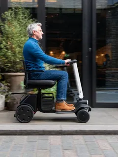 A grey-haired man in a blue sweater riding a compact mobility scooter past a cafe on a city street
