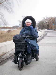 A smiling woman in a winter coat riding a rented mobility scooter along a riverside promenade